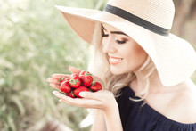 Smiling Blonde Woman 24-26 Year Old Holding Strawberry Outdoors. Wearing Straw Hat. Healthy Eating. 20s.