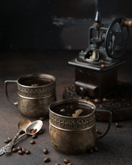 Two old vintage cups of coffee with cocoa beans, old coffee grinder and old spoons on dark brown surface opposite the dark brick wall. Still life. Coffee break