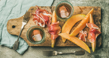 Poster - Summer wine party set. Flatlay of Wine snack Prosciutto ham, melon and glasses of rose wine with ice cubes on rustic wooden board over grey concrete background. Top view, copy space