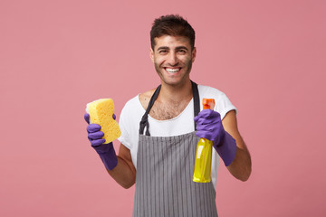 Waist-up portrait of happy positive young man with beard smiling broadly while doing housework by himself, wearing apron and protective rubber gloves, holding cleaning spray and yellow sponge