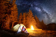 Male tourist enjoying in his camp near the forest at night. Man sitting near campfire and tent under beautiful night sky full of stars and milky way. Astrophotography