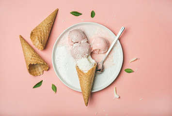 Flatlay of pastel pink strawberry and coconut ice cream scoops, sweet waffle cones on white plate over pastel pink background, top view