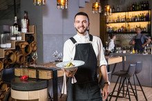 A Handsome Young Guy With A Beard Dressed In An Apron Standing In A Restaurant And Holding A White Plate With A Moth. Against The Background, The Bar Counter And Loft Style Interior