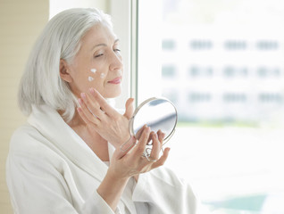 Beautiful elderly woman holding mirror and applying face cream at home