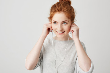 Wall Mural - Portrait of young cheerful redhead girl smiling putting on headphones over white background.