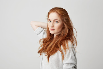 Wall Mural - Portrait of young pretty ginger girl with freckles looking at camera touching hair over white background.