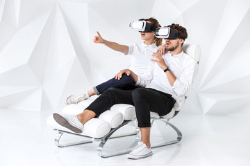 A young couple wearing VR headsets sitting on white chair in a room with white walls and floors