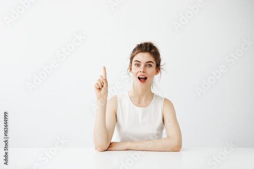Fotografie, Obraz Surprised cheerful pretty girl looking at camera with opened mouth pointing finger up sitting at table over white background