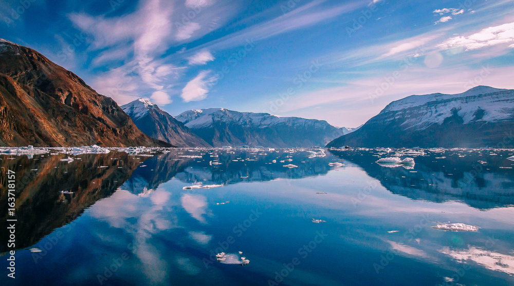 Clouds reflected in blue water of Greenland fjord surrounded by snow capped peaks. - obrazy, fototapety, plakaty 