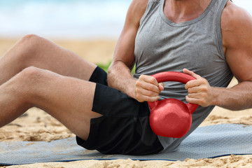 Wall Mural - Fitness man lifting kettlebell weight training russian twist exercise. Exercising on beach training with kettlebells working out core, obliques and abdominal abs muscles working out six pack.