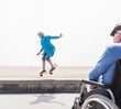 © Connect Images - Senior man in wheelchair watching wife doing skateboard trick at beach, Santa Monica, California, USA