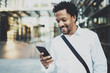 Smiling African American while walking at the city holding mobile phone in hands and checking email.Blurred background.