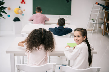 Wall Mural - Back view of pupils sitting at desks and cute schoolgirl smiling at camera in classroom