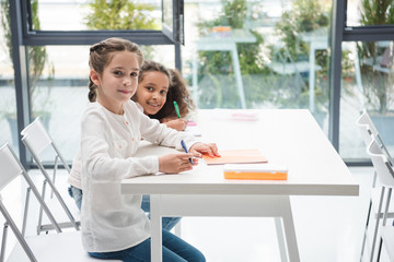 Wall Mural - Adorable multiethnic schoolgirls sitting at desk and smiling at camera in classroom