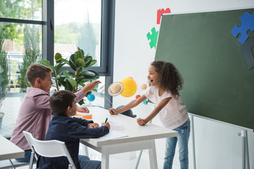Wall Mural - Adorable multiethnic schoolchildren talking and smiling at desk in classroom
