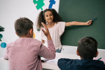 Wall Mural - Back view of schoolboy raising hand and looking at smiling african american schoolgirl pointing at chalkboard