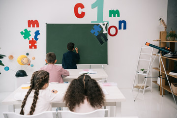 Wall Mural - Back view of pupils sitting at desks and looking at classmate writing on chalkboard