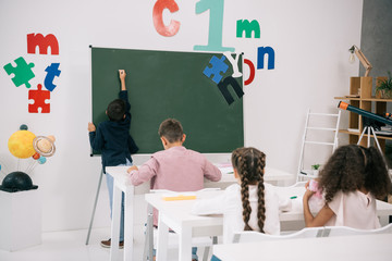 Wall Mural - Back view of schoolboy writing on chalkboard while classmates studying at desks