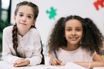 Wall Mural - Adorable multiethnic schoolgirls smiling at camera while studying together in classroom
