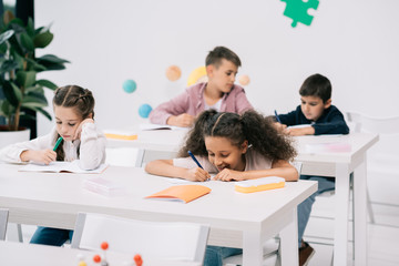Wall Mural - Multiethnic group of schoolkids writing in exercise books and studying together in classroom