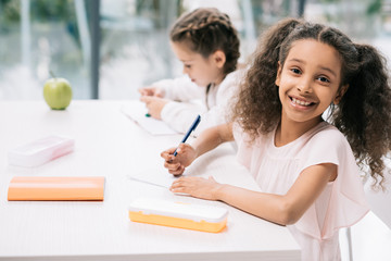 Wall Mural - Adorable african american schoolgirl smiling at camera while classmate studying behind in class