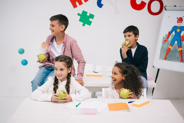 Wall Mural - Multiethnic group of children eating apples while sitting in classroom during break