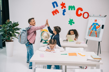 Wall Mural - Cheerful schoolboys giving high five while schoolgirls studying in classroom