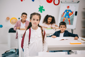 Wall Mural - Cute smiling schoolgirl with multiethnic classmates looking at camera in classroom