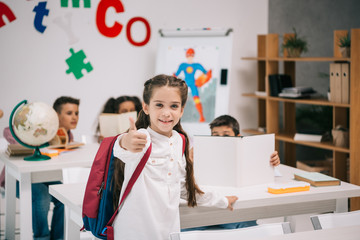 Wall Mural - Cute smiling schoolgirl showing thumb up while classmates studying behind