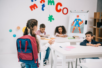 Wall Mural - Back view of new pupil with backpack looking at classmates sitting at desks
