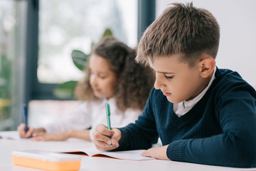 Wall Mural - Concentrated schoolboy sitting at desk and writing in exercise book with classmate sitting behind