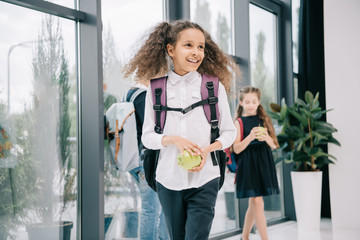 Wall Mural - Smiling african american schoolgirl holding apple and looking away in school hallway