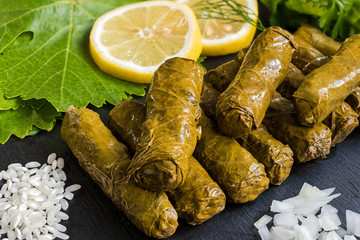 Delicious stuffed grape leaves (the traditional dolma of the mediterranean cuisine) on black dish with leaves, lemon slices, rice, parsley, dill and onion