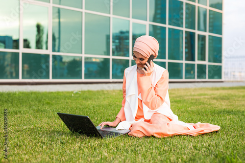 Plakat Młoda Muzułmańska kobieta Używa telefon komórkowego i laptop W parku