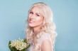 Closeup portrait of young beautiful blonde bride in spring style with bouquet of lily of the valley flowers on blue background. Wedding makeup and hairstyle in natural style. Just married