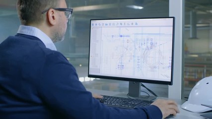 Male Industrial Engineer Works on His Computer with Technological Blueprints. Inside of the Factory is Seen From Her Office Window. Shot on RED EPIC-W 8K Helium Cinema Camera.