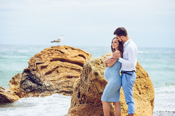 A beautiful pregnant woman in a blue dress and her courageous and handsome husband in a shirt and blue trousers leaned to each other against the background of the rock with seagull and the sea.