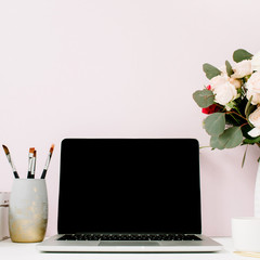Home office desk with blank screen laptop, beautiful roses and eucalyptus bouquet, white vintage casket in front of pale pastel pink background. Blog, website or social media concept .