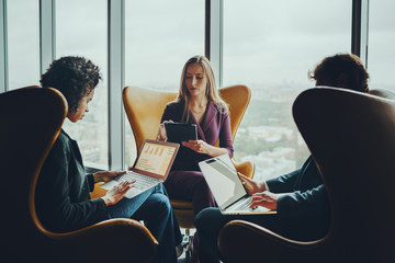 Wall Mural - Silhouettes of beautiful serious businesswoman and her coworkers sitting on yellow armchairs next to huge panoramic windows of office skyscrapers and having business meeting using their gadgets