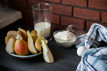 The process of preparation in detail. Cooking, kneading dough. A glass of milk, 2 eggs, a saucer with flour, a dish with pears, a whisk for whipping and a blue napkin on a black wooden background.