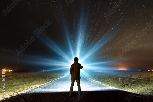 Fotografia, Obraz Young man shining a powerful torch on a starry night in Iceland, looking for the