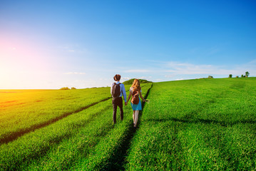 With a backpack, a man in a hat and a woman with long hair go along the path. A couple walks along the meadow