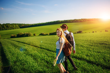 With a backpack, a man in a hat and a woman with long hair go along the path. A couple walks along the meadow