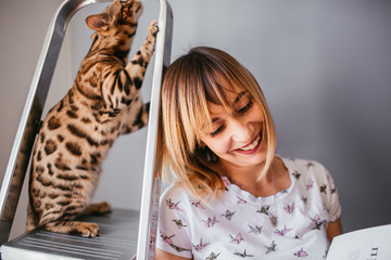 Wall Mural - Woman reads a book while Bengal cat stands on the ladder behind her