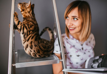 Wall Mural - Woman reads a book while Bengal cat stands on the ladder behind her