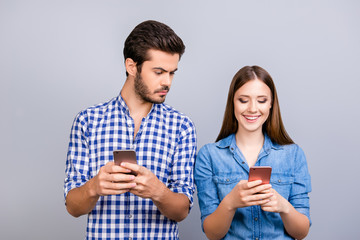 Curious boyfriend is spying his lover`s smartphone. They are wearing casual shirts, standing isolated on pure background