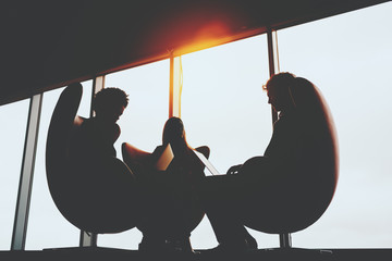 Wall Mural - Silhouettes of three business counterparts sitting on curved armchairs in front each other near office panoramic windows and having transactional meeting using their modern laptops