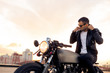 © Mikalai Bachkou - Handsome rider man with beard and mustache in black fashion sunglasses smoking cigaret and correct biker jacket sit on classic style cafe racer motorbike at sunset. Brutal fun urban lifestyle.