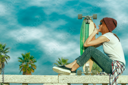 Plakat skater dziewczyna w dżinsach siedzi i trzymając deskorolka w słoneczny dzień z pochmurnego nieba. głębokie błękitne niebo i nasycone kolory
