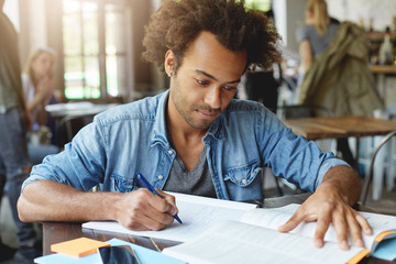 Wall Mural - Portrait of smart hardworking ambitious Afro American student sitting at university canteen, working on research, studying textbook in front of him and writing down important information in copybook